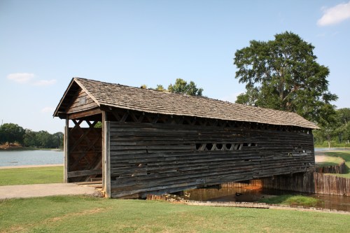 A rustic wooden covered bridge over a small stream, surrounded by green grass and trees under a clear blue sky.