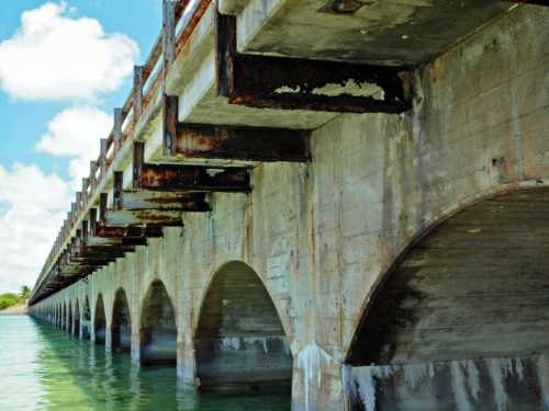 A weathered concrete bridge with arches, partially submerged in water, under a bright blue sky with scattered clouds.