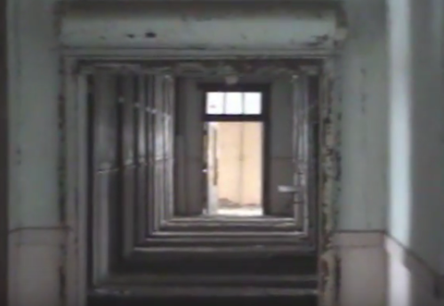 A long, dimly lit corridor with peeling walls and multiple doorways leading to a distant exit.