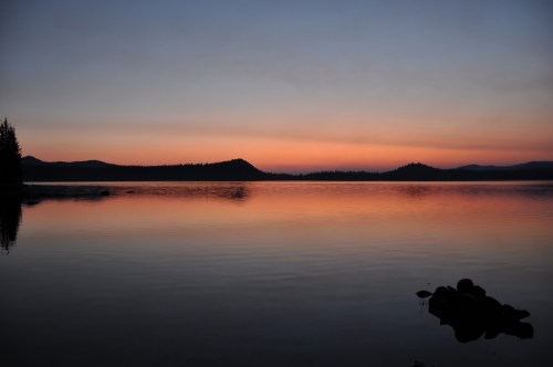 Serene lake at sunset, reflecting soft hues of orange and purple, surrounded by silhouettes of distant mountains and trees.