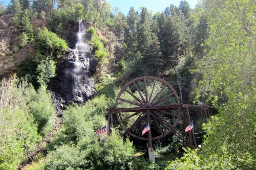 A large wooden water wheel surrounded by lush greenery and a waterfall in the background, with American flags displayed.