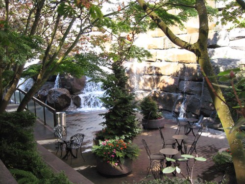 A serene outdoor space with a waterfall, surrounded by trees and tables with chairs, creating a peaceful atmosphere.
