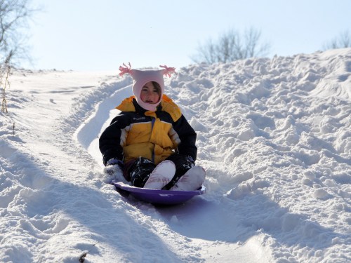 A child in a yellow jacket and pink hat sleds down a snowy hill on a sunny day.