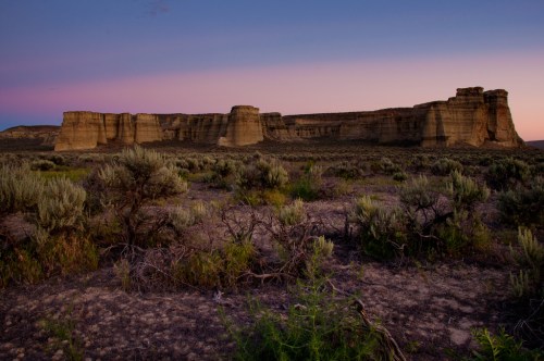 A desert landscape at sunset featuring rugged rock formations and sparse vegetation under a colorful sky.