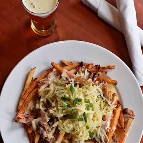 A plate of loaded fries topped with cheese, green onions, and pulled meat, alongside a small glass of beer.