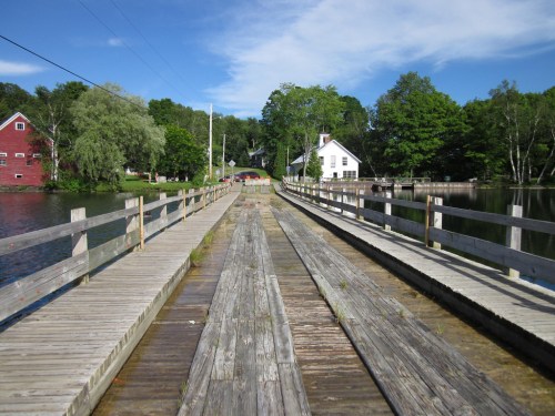 A wooden walkway extends over calm water, flanked by trees and buildings under a clear blue sky.