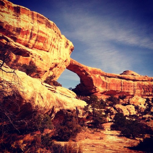 A natural rock arch stands tall against a blue sky, surrounded by rugged terrain and sparse vegetation.