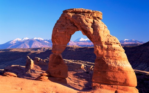 A natural stone arch stands against a clear blue sky, with snow-capped mountains in the background.