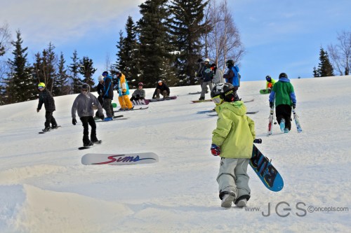 A group of snowboarders and skiers on a snowy slope, with trees in the background and a clear blue sky.