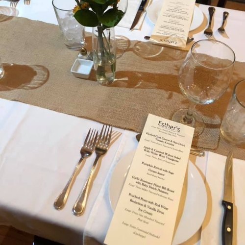 Table setting with a menu, glassware, and a small vase of flowers on a burlap runner.