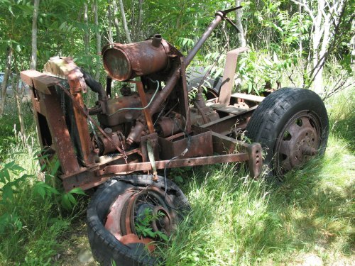 An old, rusted tractor frame sits overgrown with grass and plants in a wooded area.