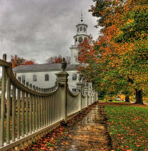 A white church with a clock tower surrounded by colorful autumn trees and a wet pathway lined by a white fence.