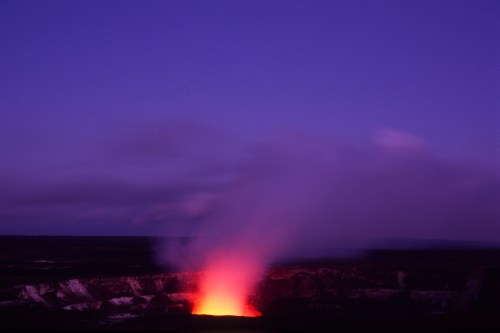 A glowing volcano erupts at dusk, with bright orange lava and smoke against a deep blue sky.