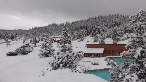 A snowy landscape with a lodge, parked vehicles, and trees, under a cloudy sky.
