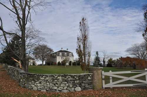 A large house behind a stone wall, surrounded by trees and a grassy area under a cloudy sky.