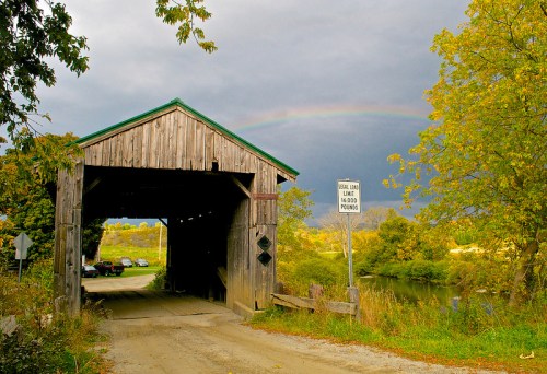 A rustic covered bridge under a cloudy sky, with a rainbow and lush greenery surrounding the entrance.