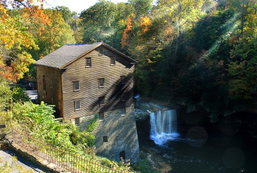 A historic wooden mill beside a waterfall, surrounded by colorful autumn foliage and a tranquil river.