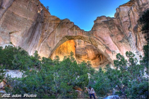 A person stands in a lush area beneath a large rock arch, surrounded by towering cliffs and a clear blue sky.