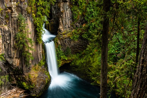 A serene waterfall cascades into a lush green forest, surrounded by rocky cliffs and vibrant foliage.