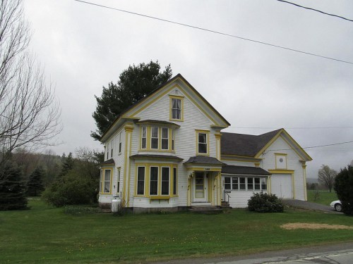 A two-story white house with yellow trim, surrounded by grass and trees, under a cloudy sky.