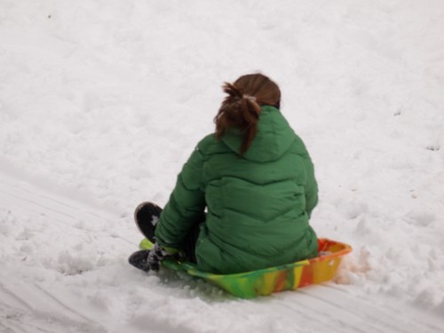 A child in a green jacket sits on a colorful sled, ready to go down a snowy hill.