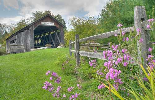 A rustic covered bridge surrounded by greenery and colorful flowers, with a wooden fence in the foreground.