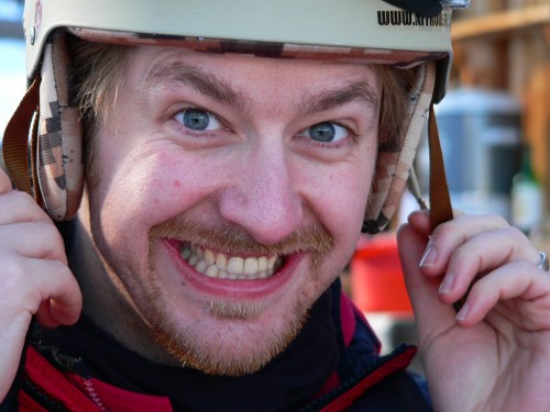A smiling man wearing a helmet, holding the straps, with bright blue eyes and a cheerful expression.