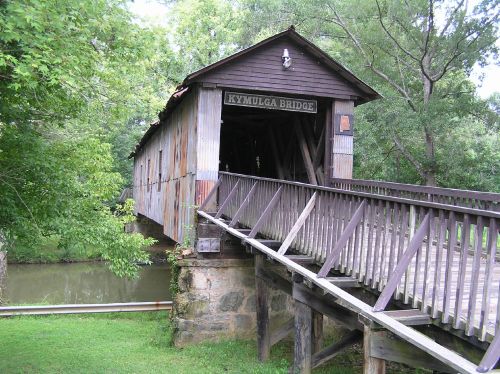 A rustic covered bridge with a wooden walkway, surrounded by lush greenery and reflecting in the water below.