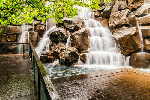A serene waterfall cascading over large rocks, surrounded by lush greenery and a stone pathway.