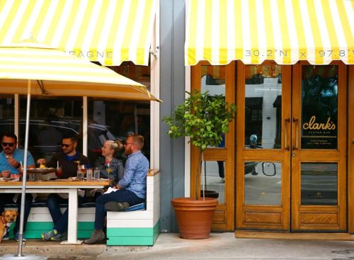 A group of people dining outside a restaurant with yellow-striped awnings and wooden doors. A dog sits nearby.