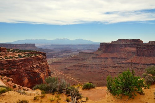 A vast desert landscape with layered rock formations and distant snow-capped mountains under a cloudy sky.