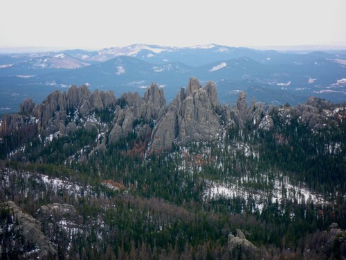 A panoramic view of rugged mountains and rocky formations, surrounded by evergreen trees and patches of snow.
