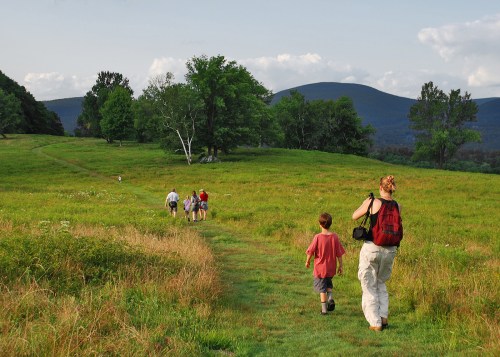 A woman and child walk along a grassy path, leading a group through a scenic landscape with mountains in the background.