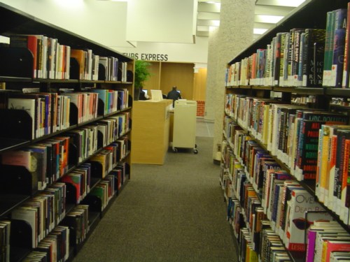 A view of library shelves filled with books, leading to a service desk in the background.