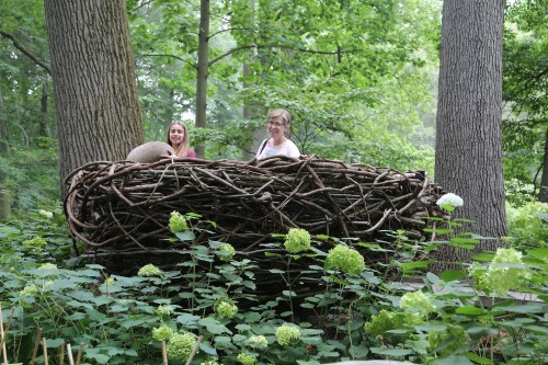Two women stand inside a large, woven nest structure surrounded by lush greenery and trees.