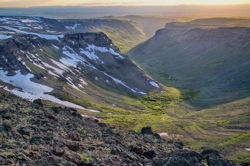 A scenic valley with rocky cliffs, snow patches, and lush greenery illuminated by soft sunlight at sunset.