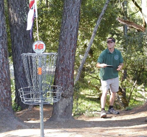 A man in a green shirt prepares to throw a disc towards a disc golf basket surrounded by trees.