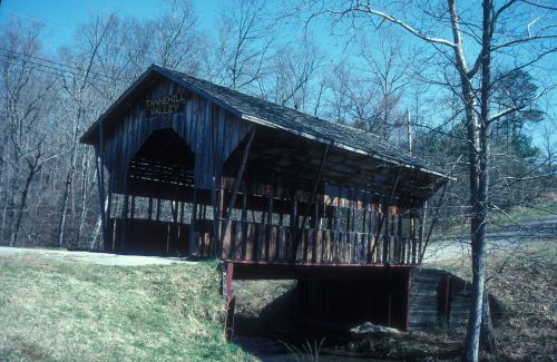A rustic wooden covered bridge stands over a small stream, surrounded by bare trees and grassy areas.