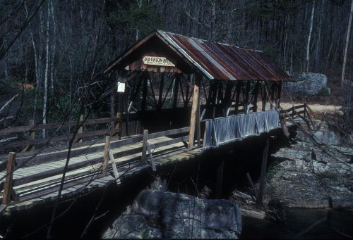A rustic covered bridge with a wooden walkway, surrounded by trees and rocky terrain.