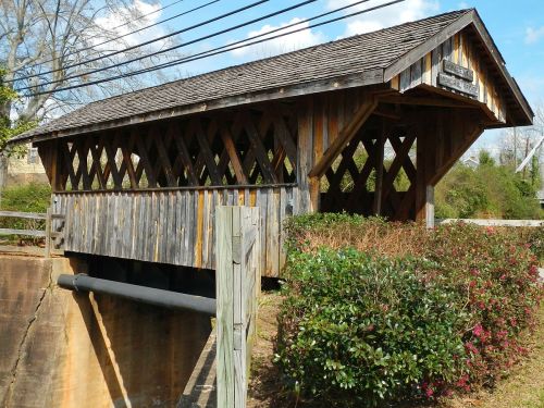 A wooden covered bridge with a sloped roof, surrounded by greenery and power lines under a blue sky.