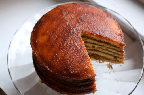 A round layered cake with a slice cut out, displayed on a glass plate. The cake has a golden-brown exterior.