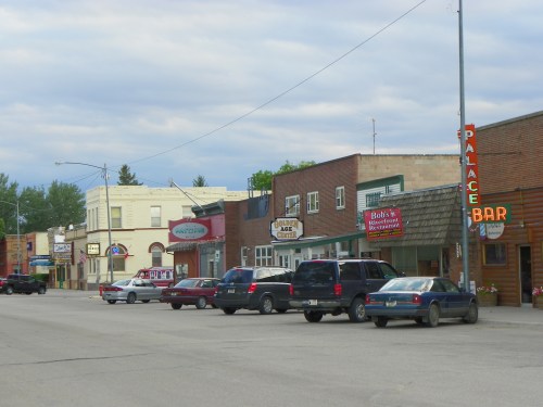 A small town street lined with shops and bars, featuring parked cars and a cloudy sky overhead.