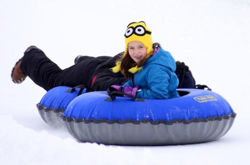 A child in a blue jacket and yellow hat smiles while lying on a blue snow tube in a snowy landscape.
