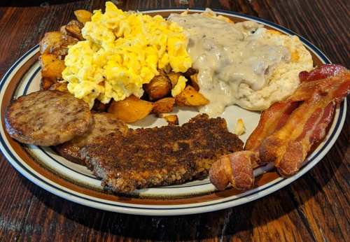 A hearty breakfast plate with scrambled eggs, sausage, bacon, potatoes, and biscuits with gravy.