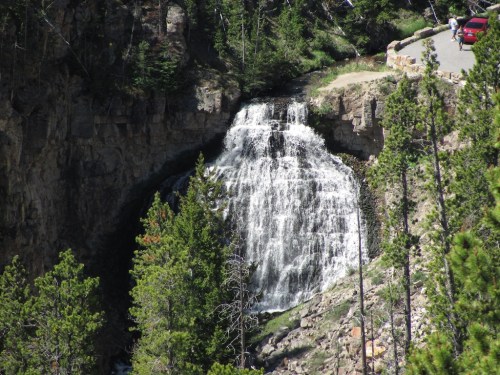 A waterfall cascades down rocky cliffs, surrounded by lush green trees, with a road and a car visible nearby.