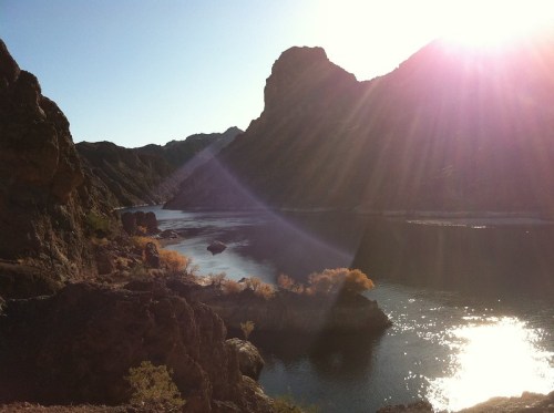 Sunlight beams over a serene river surrounded by rocky mountains and autumn foliage.
