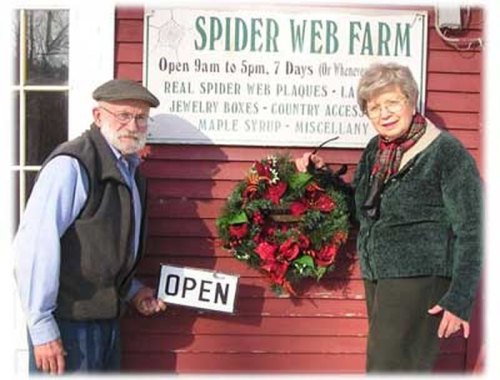 An elderly man and woman stand outside Spider Web Farm, holding an "OPEN" sign and a festive wreath.
