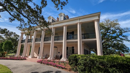 A large, elegant two-story house with columns, surrounded by greenery and flowers under a clear blue sky.