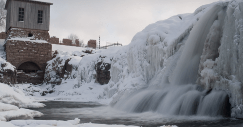 A frozen waterfall cascades over icy rocks, with a historic building in the background and snow covering the landscape.