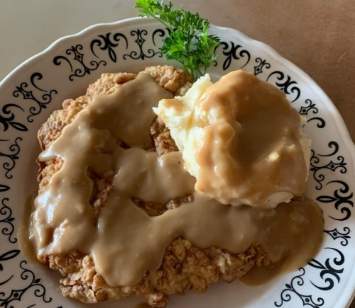 A plate of chicken-fried steak topped with brown gravy, served with a side of mashed potatoes and a sprig of parsley.